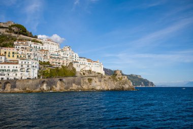 Parlak Akdeniz ışığı, İtalya 'nın Amalfi kentindeki, derin mavi suların üzerindeki sarp kayalıklara tünemiş beyaz ve pastel evleri aydınlatıyor. Dramatik kıyı şeridinde tarihi taş kemerler ve canlı bir gökyüzüne karşı kurulmuş bir gözetleme kulesi yer alıyor..