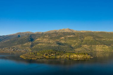 Ioannina Gölü 'ndeki ağaçlarla kaplı adanın geniş hava manzarası Pindus Dağları' nın arka planında canlı, mavi bir gökyüzünün altında. Sakin su ve bereketli yeşillik Yunanistan 'ın Epirus kentinin doğal manzarasını vurguluyor.