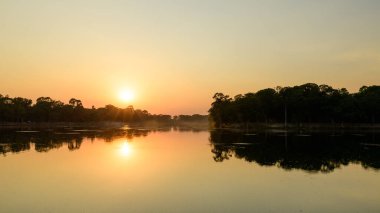 Güneş, Kamboçya 'nın Siem Reap kentindeki Angkor tapınağı kompleksi yakınlarındaki yoğun tropikal ormanlarla çevrili sakin bir göl üzerinde yükselirken, yumuşak altın ışık gökyüzünü doldurur. Durgun su, ağaçların siluetlerini ve şafağın ılık tonlarını yansıtır.