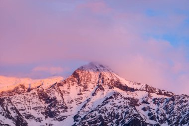 Yumuşak pembe ve mor bulutlar Aiguille du Gouter 'ın karla kaplı zirvesinde Fransız Alpleri' nin engebeli yamaçlarını aydınlatan yumuşak alpenglasla sürükleniyor..