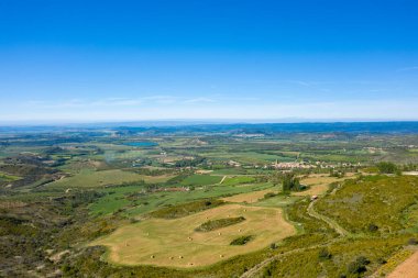 Saman balyaları ve İspanya 'nın Loarre yakınlarındaki uzak bir köy ile noktalanmış yeşil tarlaların geniş hava manzarası. Kırsal bölgeler parlak mavi bir gökyüzü altında uzanır, dalgalı tepeler ve parlak güneş ışığında açık tarım alanları sergilenir..