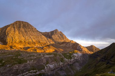 Altın akşam güneşi, çizgili kaya yüzlerini ve Pireneler 'deki dramatik bir dağ yığınının dik yamaçlarını aydınlatır. Karşıt gölgeler ve karamsar bir gökyüzü vardır..