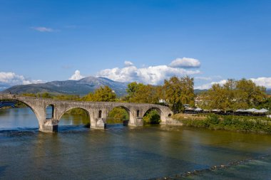 Yunanistan 'ın Arta kentindeki tarihi kemerli taş köprünün geniş manzarası, uzak dağlar ve parlak mavi gökyüzünün altındaki sonbahar ağaçlarıyla temiz bir nehri geçiyor. Nehir kenarındaki kafelerde şemsiyeler ve yemyeşil alanlar. Nehir kenarında hareketli bir yolculuk yapıyorlar.