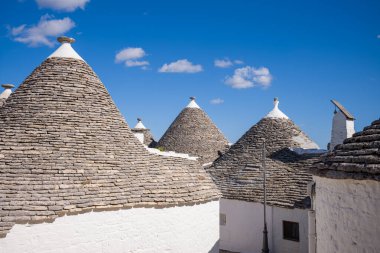 Bir grup geleneksel trullo evi konik taştan çatıları ve beyazlatılmış duvarları olan Alberobello, Apulia, İtalya 'da parlak mavi bir gökyüzünün altında duruyor. Sahne kireçtaşı yapısı ve belirgin Akdeniz mimarisini vurguluyor