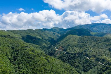 Laos 'un başkenti Phou Khoun ile Luang Prabang arasında, sık yeşil tepeler ve vadilerden geçen dolambaçlı bir dağ yolunun hava manzarası. Manzara canlı bitki örtüsü, katmanlı tepeler ve dramatik bulut oluşumları içerir.