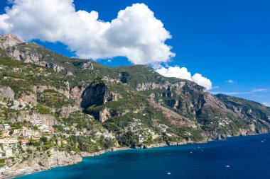Dik kireçtaşı dağları, İtalya 'nın Positano yakınlarındaki Amalfi Sahili' nin canlı mavi sularının üzerinde yükseliyor. Dağınık evler ve yemyeşil Akdeniz bitkileri parlak bir gökyüzünün altında dalgalı bulutlarla dolu dramatik kıyı yamaçlarını noktalıyor..