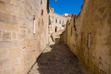 Matera, İtalya 'da güneşli bir sokak. Kireçtaşı duvarları ve kaldırım taşlarıyla kaplı. Sahnede sıcak ışık, derin gölgeler ve sakin bir Akdeniz atmosferi yer alıyor..