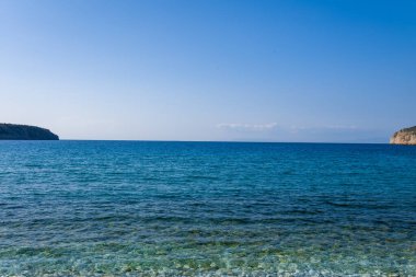 Şeffaf mavi suyun sakin manzarası çakıl taşlı bir plajla buluşuyor, uzak kayalık burunlar ve bulutsuz bir gökyüzü sakin bir sahil manzarası yaratıyor..