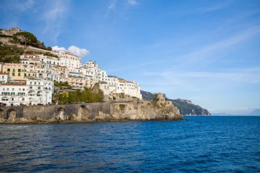 İtalya 'nın Amalfi kentindeki sarp kayalıklar boyunca uzanan güneşli beyaz ve pastel binalar, derin mavi Akdeniz' e bakıyor. Dramatik kıyı şeridinde tarihi taş kemerler ve açık gökyüzünün altında bir ortaçağ gözlem kulesi yer alıyor..