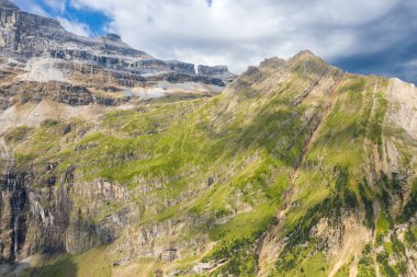 Yeşil çimenli yamaçları olan dik, çizgili dağ uçurumları ve Pireneler 'deki dramatik kaya katmanları parçalı bulutlu bir gökyüzünün altında yer değiştiren güneş ışığıyla aydınlatılıyor. Engebeli dokular ve keskin jeolojik çizgiler alp arazisine hakim..