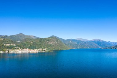 Kuzey İtalya 'daki Orta Gölü' nün geniş bir manzarası. Ormanlık tepelerle çevrili derin mavi sular ve berrak bir gökyüzünün altındaki uzak dağ zirveleri. Sahil şeridi küçük köylerle dolu. Huzurlu bir göl manzarası yaratıyor..