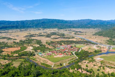Geniş hava perspektifi, kırmızı çatılı binaların ekili tarlalar, dolambaçlı nehirler ve açık düzlüklerle çevrili olduğu küçük bir yerleşim yeri gösteriyor. Manzara, ormanlık tepelerle ve açık mavi gökyüzünün altındaki uzak dağlarla sınırlıdır.