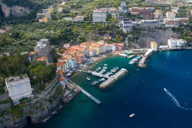 İtalya 'nın Sorrento kentinin yüksek açılı perspektifi, rıhtım boyunca kenetlenmiş tekneleri ve dramatik uçurumların altında yığılmış pastel binaları olan canlı bir marinayı gösteriyor. Sahnede yemyeşil, berrak mavi su ve canlı bir Akdeniz kıyısı yer alıyor.