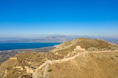 Akrokorinth 'in güneşli yamaçlarında süzülen antik taş tahkimatlarının hava görüntüsü, canlı mavi su ve uzak dağlar Yunanistan' da çarpıcı bir zemin oluşturuyor..
