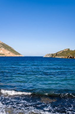 Yunanistan 'ın Porto Kagio kentindeki engebeli burunlar arasında uzanan berrak mavi su, canlı bir gökyüzünün altındaki taştan bir kıyıda hafif dalgaların kırılmasıyla birlikte. Sahne parlak ve sakin, doğal kıyı dokusunu vurguluyor..