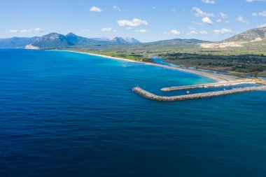 Orosei marina iskelesinin hava manzarası canlı mavi Akdeniz sularına doğru kıvrılıyor, uzun kumlu kıyı şeridi ve parlak bir gökyüzünün altındaki uzak Sardunya dağları.