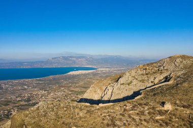 Akrokorinth 'in kayalık yamaçlarından geniş bir arazi, mavi körfeze, uzak dağlara ve berrak bir gökyüzünün altındaki yamalı tarlalara bakıyor..