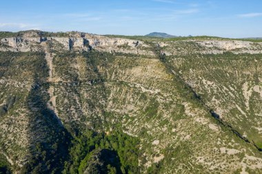 Navacelles Sirki 'nin sarp, yeşil kaplı kanyonuna bakan engebeli bir kireçtaşı platosunun yüksek görüş açısı. Parlak güneş ışığı açık mavi gökyüzünün altındaki kayalık kayalıkları ve dokulu yamaçları aydınlatır..
