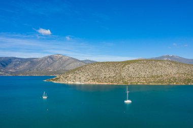 İki yelkenli, kayalık, çalılarla kaplı tepeler ve berrak mavi bir gökyüzünün altındaki uzak dağlarla çevrili korunaklı bir körfezde turkuaz sularda yüzüyor. Parlak güneş ışığı, sakin ve davetkar Akdeniz manzarasını güçlendirir.