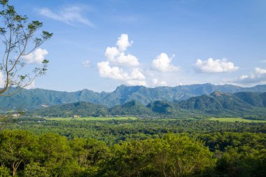 Yoğun orman ve açık arazilerle dolu yemyeşil bir vadinin engebeli mavi dağlarla çevrili parlak bir gökyüzü altında ve Kuzey Vietnam 'da dağınık bulutlar var. Manzara geniş, güneşli ve huzurlu görünüyor..