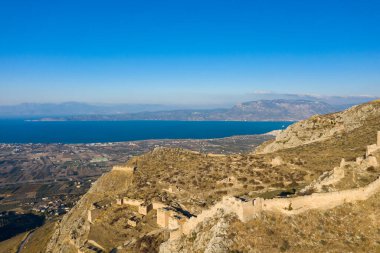 Antik kale, Akrokorint 'in engebeli yamaçları boyunca uzanmaya devam ediyor, Yunanistan' da berrak bir gökyüzü altında canlı mavi Corinthian Körfezi ve tarım arazilerine tepeden bakan.