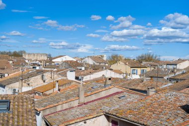 Aigues-Mortes 'in tarihi merkezinde yıpranmış terracotta çatıları üzerinde geniş bir manzara, desenli fayanslar ve parlak bacalar dağınık mavi bulutlarla dolu canlı bir gökyüzüne karşı kurulmuş..