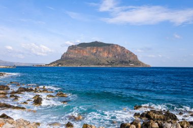 Monemvasias 'ın sarp kayalık adası canlı mavi Akdeniz suları üzerinde yükseliyor, kıyı şeridi kayaları ve parlak, parçalı bulutlu gökyüzü altında hafif dalgalar.
