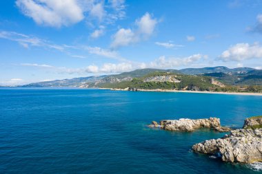 Yunanistan 'ın Epirus kentindeki Karavostasi körfezinin geniş bir manzarasında, turkuaz su ve kumlu bir kıyı şeridinin üzerindeki ormanlık bir tepenin üzerinde çağdaş bir tatil beldesi yer alıyor. Parlak gün ışığı ve dağınık bulutlar Akdeniz kıyı manzarasını ve engebeli araziyi vurgular.