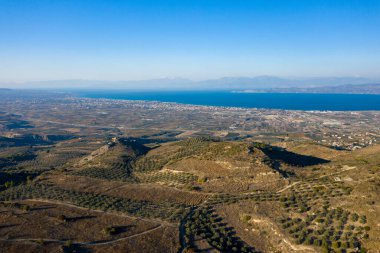 Yuvarlanan zeytin korulukları, açık tarım arazileri ve berrak bir gökyüzünün altında canlı mavi bir deniz içeren havadan manzaralar. Sahne, ufku çevreleyen uzak dağlarla güneşli ve geniştir..