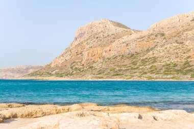 Parlak turkuaz su, Yunanistan 'ın Girit kentindeki Balos Sahili' ndeki engebeli güneşli dağlarla buluşuyor. Açık gökyüzü ve dokulu kayalar, canlı ve açık bir kıyı manzarası yaratıyor..