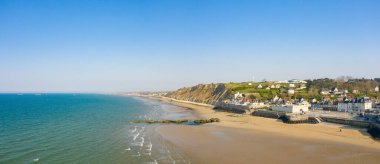 Kumlu sahil manzarası, turkuaz deniz ve Arromanches-les-Bains 'teki sarp çimenli uçurumlara karşı kurulmuş büyüleyici bir köy. Açık gökyüzü ve yumuşak güneş ışığı sakin ve davetkar bir atmosfer yaratır..