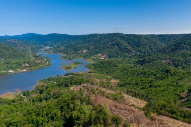 Hava perspektifi, yemyeşil tepeler ve açık mavi gökyüzünün altındaki ormansız arazi bölgeleri arasında dolaşan bir nehri yakalar. Manzara yoğun bitki örtüsü, korunmasız toprak ve doğal eğriler içeriyor.