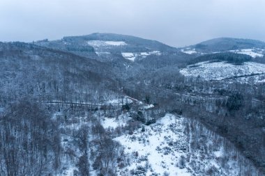 Karla kaplı tepelerin ve Morvan bölgesindeki yoğun kış ormanlarının havadan görünüşü dağınık açıklıklar ve sessiz mavi bulutlu gökyüzü sakin ve uzak bir atmosfer yaratıyor..