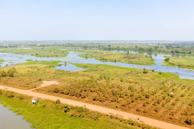 Kamboçya 'nın Phnom Penh kentinin eteklerinde uzanan dağınık göletleri, çimenli tarlaları ve toprak yolu olan geniş bir kırsal alan manzarası. Parlak gün ışığı, doğal dokuları, su kanallarını ve bazı bölgeleri vurgular