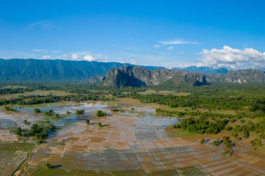 Su basmış pirinç tarlalarının, yoğun yeşil orman ve çarpıcı kireçtaşı çıkıntılarıyla çevrili kırsal bir ovada geometrik bir mozaik oluşturması. Manzara katmanlı dokuları, açık tarım arazilerini ve geniş arazileri vurguluyor.