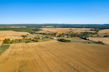 Geniş altın tarlalar, dağınık ağaçlar ve orta Fransa 'da canlı mavi bir gökyüzünün altındaki uzak ormanlık alanlarla çevrili küçük bir kırsal köyün havadan görünüşü. Açık arazi ve yamalı tarım arazisi, geniş bir alan ve kırsal alan hissi yaratır.