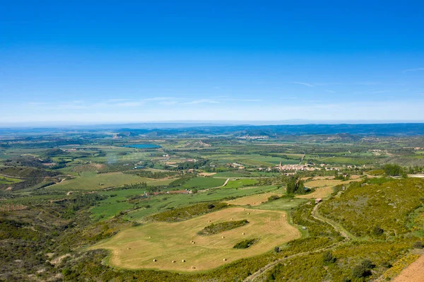 Saman balyaları ve İspanya 'nın Loarre yakınlarındaki uzak bir köy ile noktalanmış yeşil tarlaların geniş hava manzarası. Kırsal bölgeler parlak mavi bir gökyüzü altında uzanır, dalgalı tepeler ve parlak güneş ışığında açık tarım alanları sergilenir..