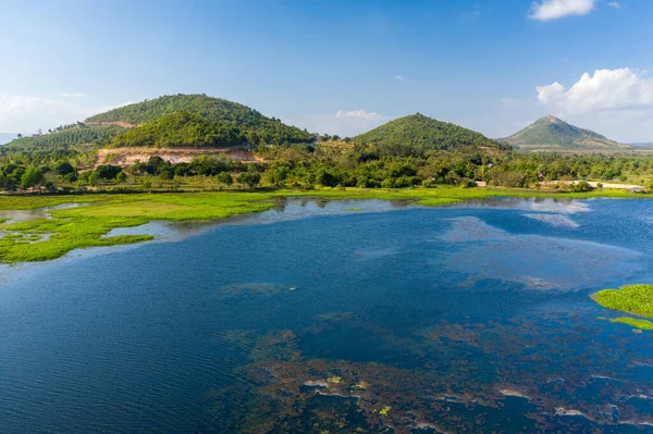 Kamboçya 'nın Kampong Chhnang kentindeki Brateak Krola Gölü' nün sakin mavi sularının ve su bitkilerinin ardında yemyeşil tepeler yükselir. Parlak gün ışığı kıyı şeridinin doğal dokusunu ve yukarıdaki açık gökyüzünü vurgular..