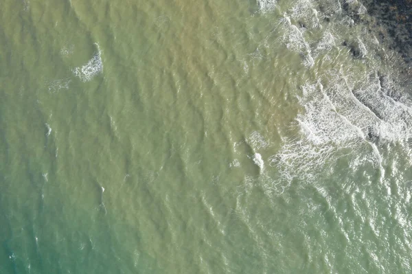 Yukarıdan aşağıya insansız hava aracı görüntüsü kıyıya yakın yeşilimsi deniz suyunda yumuşak dalgalar ve ince dalga desenleri yakalıyor. Güneş ışığı, okyanus yüzeyinin doğal dokusunu ve hareketini vurgular..