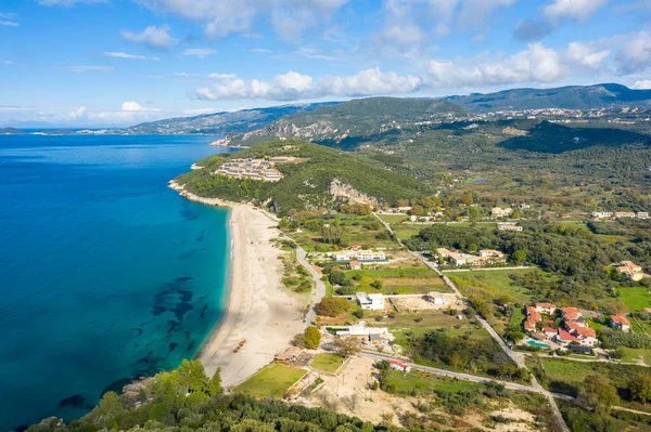 Yunanistan 'ın Epirus kentindeki Karavostasi plajının yüksek açılı manzarası, turkuaz su, geniş kumlu bir kıyı şeridi ve ormanlık bir tepenin üzerinde modern bir tatil köyü. Parlak gün ışığı ve dağınık bulutlar Akdeniz manzarasını ve uzanan yeşil tepeleri gözler önüne seriyor