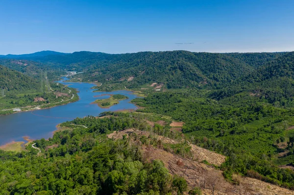 Hava perspektifi, yemyeşil tepeler ve açık mavi gökyüzünün altındaki ormansız arazi bölgeleri arasında dolaşan bir nehri yakalar. Manzara yoğun bitki örtüsü, korunmasız toprak ve doğal eğriler içeriyor.