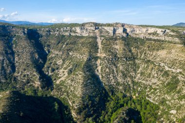 Navacelles Sirki 'nde yoğun yeşil bitki örtüsüyle dolu derin bir kanyonun üzerinde dramatik kireçtaşı kayalıkları yükselir. Güneş ışığı engebeli taş dokuları aydınlatırken gölgeler sarp arazide çarpıcı bir kontrastlık yaratır..