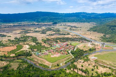 Kırmızı çatılı binalarla çevrili planlanan bir yerleşim alanı kıvrımlı bir nehrin kenarında, yama alanları, ağaçlık alanlar ve uzak tepelerle çevrili. Manzarada geometrik planlar, ekili arazi ve açık kırsal alanlar açık gün ışığı altında yer almaktadır..