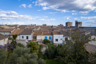 Aigues-Mortes 'in merkezinde geleneksel kiremitli çatılar ve yemyeşil bahçeler, tarihi taş duvarlar ve arka planda parlak, bulutlu bir gökyüzünün altında görünen kuleler.