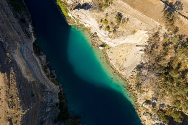Korint Kanalı 'nın yukarıdan aşağıya canlı turkuaz su, dik kayalık kıyılar ve kıyı boyunca seyrek bitki örtüsüyle güneşli kurak arazi manzarası..