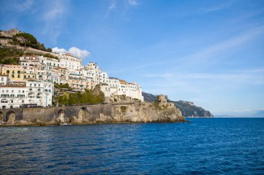 İtalya 'nın dramatik Amalfi kıyı şeridi boyunca uzanan tarihi taş kemerlerin üzerinde beyaz ve pastel binalar yığılıyor. Sahnede dik uçurumlar, bir ortaçağ gözetleme kulesi ve berrak bir gökyüzünün altındaki derin mavi Akdeniz suları yer alıyor..