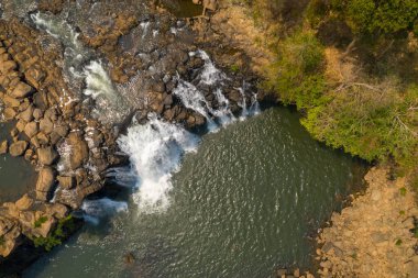 Tepetaklak bir perspektif, Tad Lo şelalesinin engebeli kayaların üzerinden akan suları, dağınık kayalar ve güney Laos 'taki seyrek yeşilliklerle çevrili sakin bir havuza yansıtıyor. Güneş ışığı, dokulu taşları ve yumuşak akışı vurgular, huzur uyandırır.