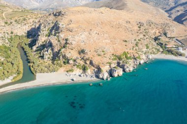 Girit 'teki Preveli plajının hava manzarasında engebeli güneşli uçurumlar, palmiye ağaçlarıyla kaplı dolambaçlı bir nehir ve kumlu bir kıyı şeridi boyunca berrak turkuaz bir su görülüyor. Kurak tepeler yemyeşil ve canlı mavi deniz ile tezat oluşturur..