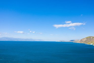 Uzak dağlara doğru uzanan derin, mavi bir denizin geniş manzarası, güneşli tepeler ve canlı, bulutsuz bir gökyüzünün altında dağınık bulutlar. Sahne açık, huzurlu ve doğal berraklıkla dolu..