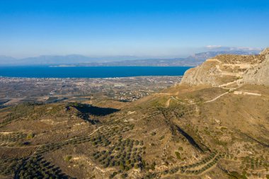 Yunanistan 'da berrak bir gökyüzünün altında geniş mavi bir uçurum ve uzak dağlara bakan engebeli bir uçurumun üzerindeki engebeli kayalıklara uzanan dağınık zeytin ağaçlarıyla teraslı tepe manzarası..